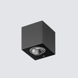 A sleek black, cubic outdoor wall light casts two cones of warm, inviting light—one upward and one downward—on a textured stone wall at night. In the background, a modern house with a dark wooden door and large windows is dimly lit under a starry sky.