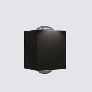 A sleek black, cubic outdoor wall light casts two cones of warm, inviting light—one upward and one downward—on a textured stone wall at night. In the background, a modern house with a dark wooden door and large windows is dimly lit under a starry sky.