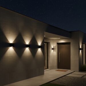 A sleek black, cubic outdoor wall light casts two cones of warm, inviting light—one upward and one downward—on a textured stone wall at night. In the background, a modern house with a dark wooden door and large windows is dimly lit under a starry sky.