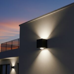 A sleek black, cubic outdoor wall light casts two cones of warm, inviting light—one upward and one downward—on a textured stone wall at night. In the background, a modern house with a dark wooden door and large windows is dimly lit under a starry sky.